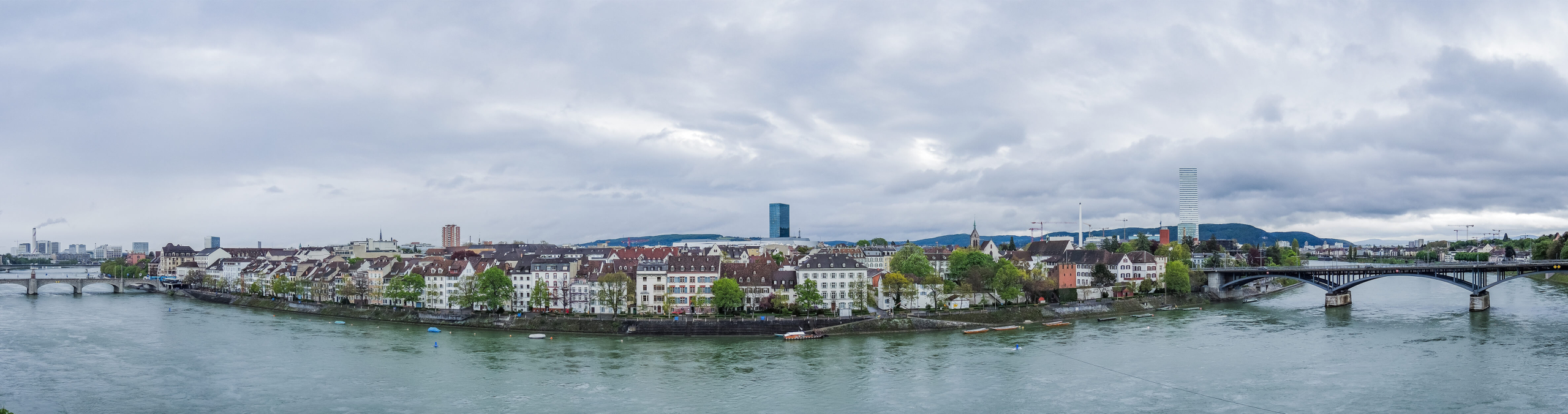 Hridgandhaa pano 4 Panoramic view of Basel's riverfront architecture and bridges under a cloudy sky.