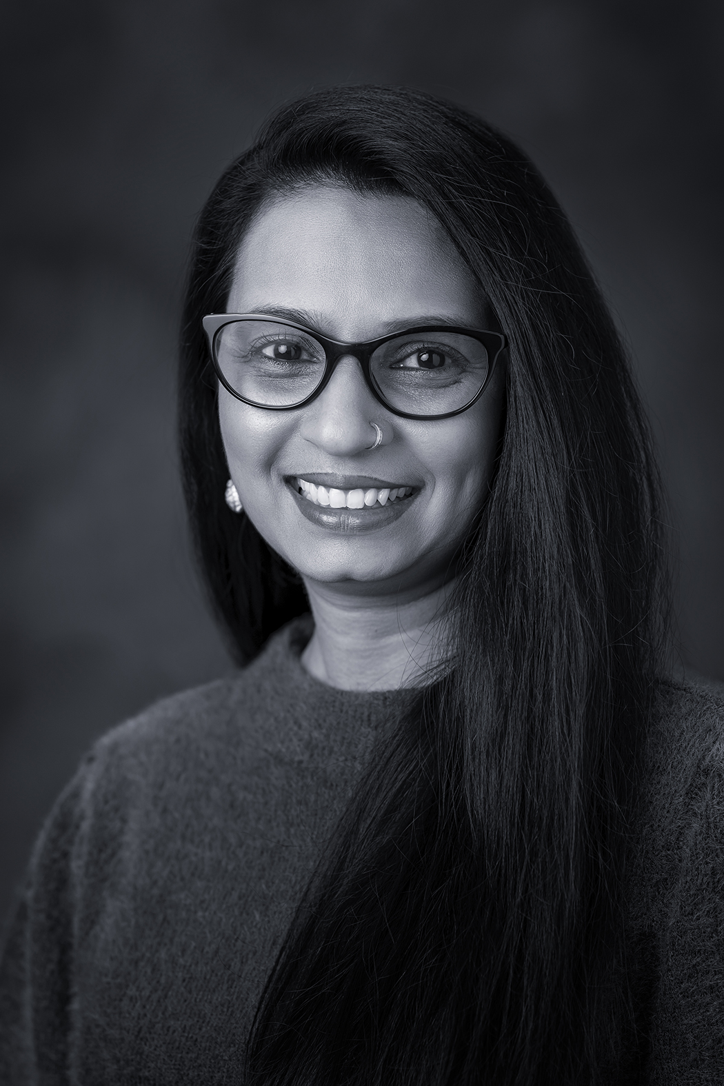 hridgandhaa Profile about her Portrait - Hridgandha Mistry smiling, wearing glasses and a dark sweater, against a dark blurred background.