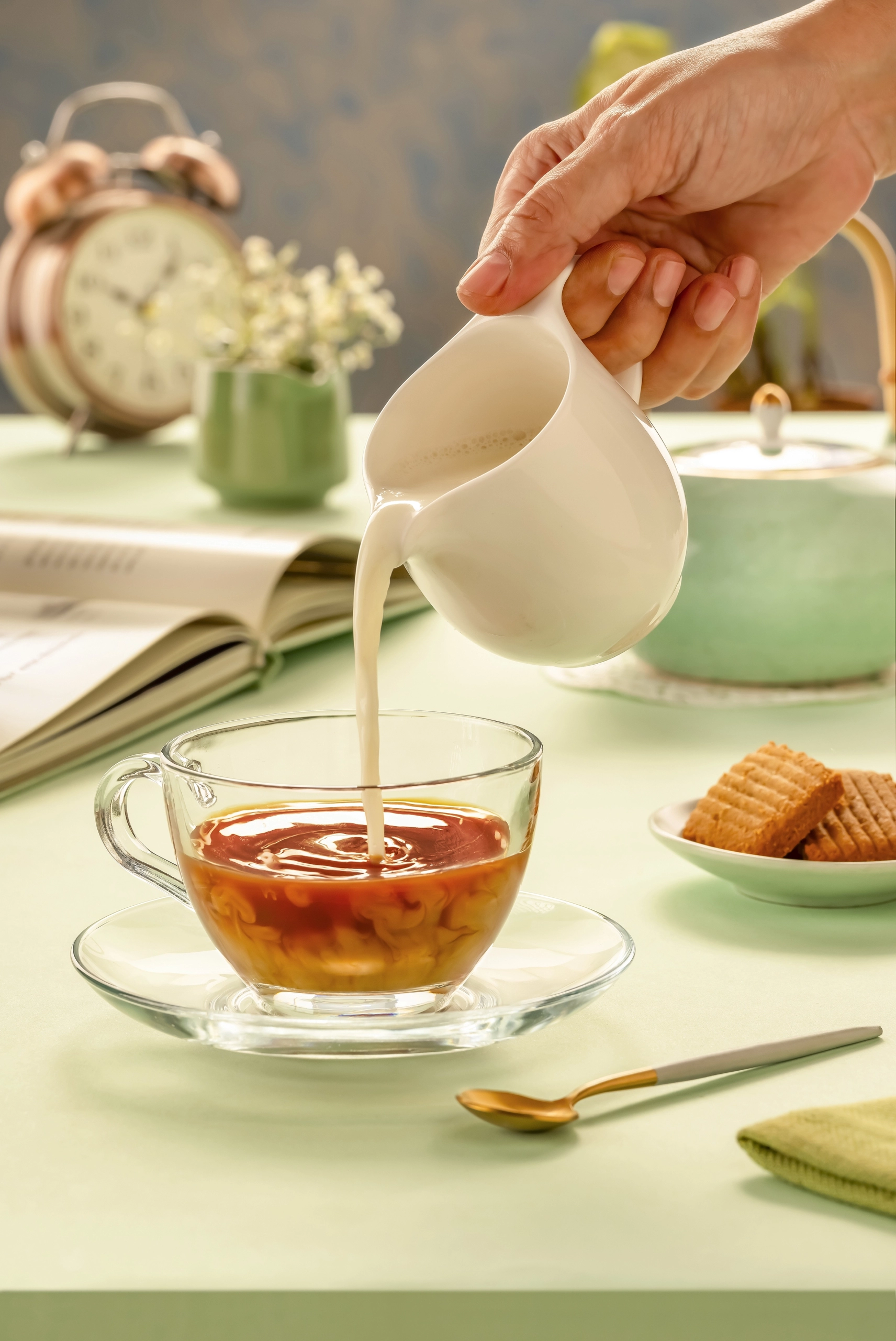 Milk being poured into a glass cup of tea on a pastel table with books and biscuits in the background.