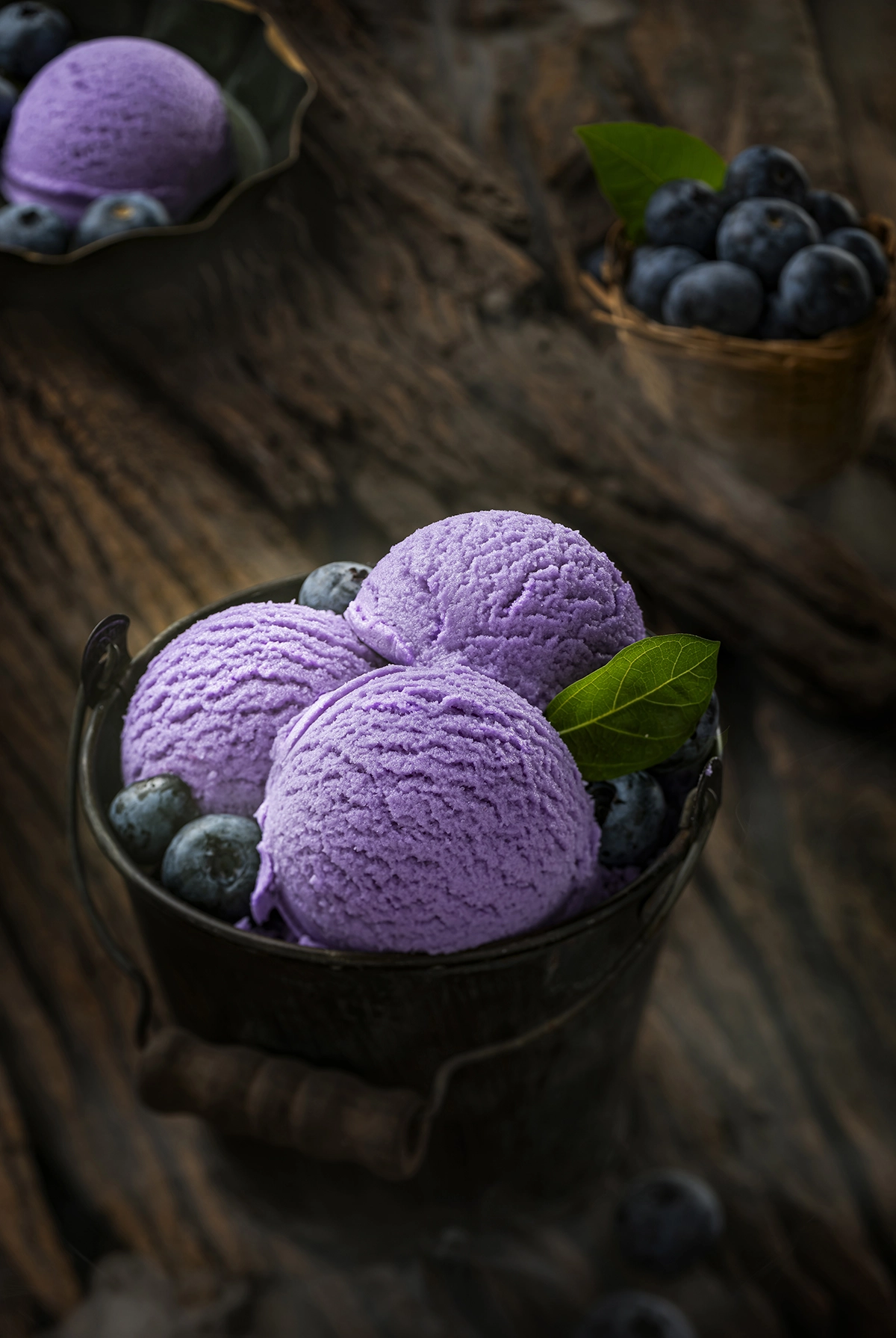 Scoops of blueberry ice cream in a dark bowl with fresh blueberries on a rustic wooden surface.